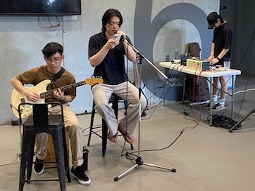 three individuals performing a simple busking performance. In the foreground of the photo, one person is on a guitar, one is singing into a microphone, and a person is in the background on a sound mixer engineering the sound.