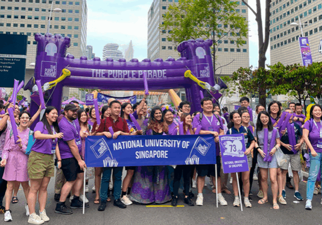A group of students wearing purple, holding a banner reading "National University of Singapore" at the Purple Parade contingent march