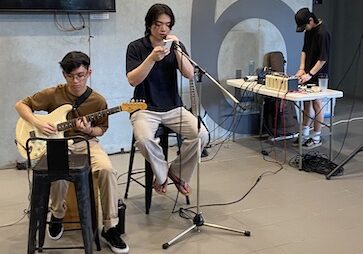 three individuals performing a simple busking performance. In the foreground of the photo, one person is on a guitar, one is singing into a microphone, and a person is in the background on a sound mixer engineering the sound.